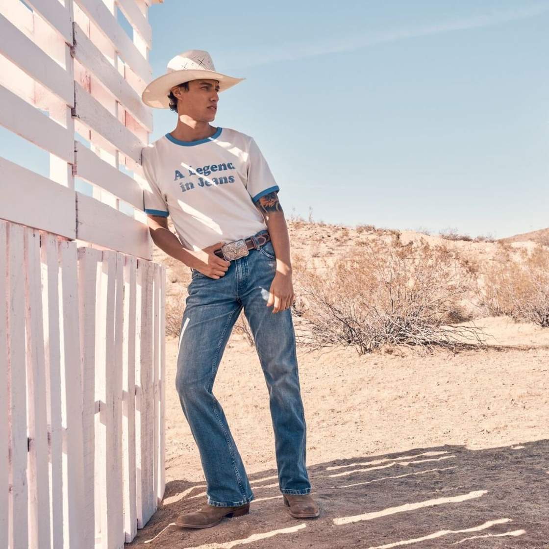 Image of a man standing in the shade, wearing Wrangler jeans, a white t-shirt with blue details that reads 'A legend in jeans'. He has a cowboy hat, boots and belt buckle.