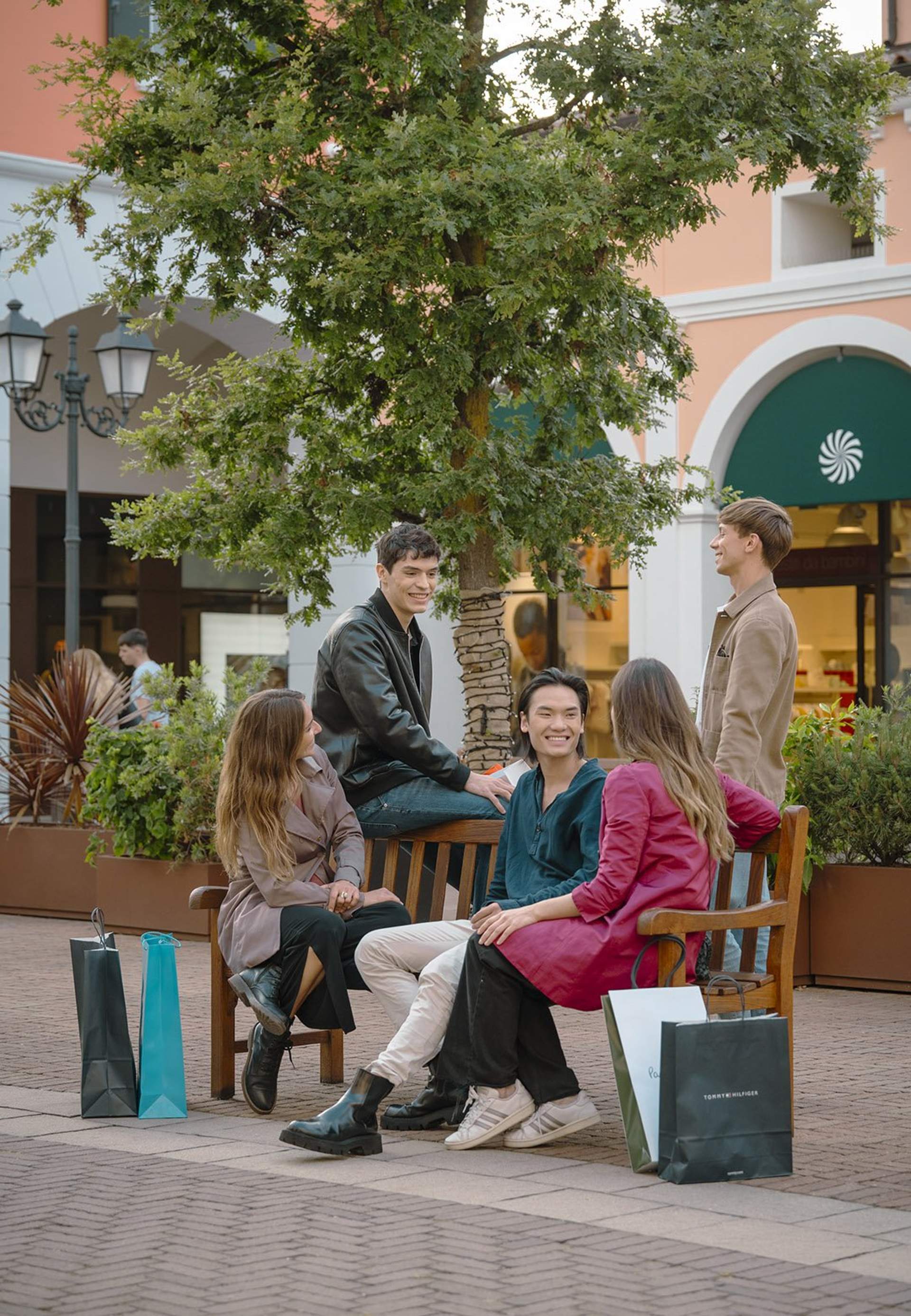 Group of friends talking on a bench at Noventa di Piave Designer Outlet with shopping bags.